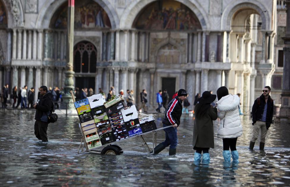 a man pulls a cart carrying fruits and vegetables through a flooded st. mark's square during a period of seasonal high water in venice
