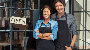 two cheerful small business owners smiling and looking at camera while standing at entrance door. happy mature man and mid woman at entrance of newly opened restaurant with open sign board. smiling couple welcoming customers to small business shop