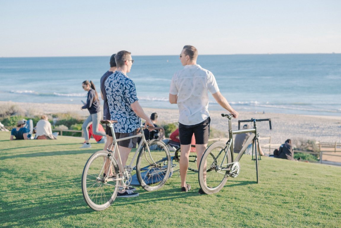 three men with commuter bikes on grass field front of sea