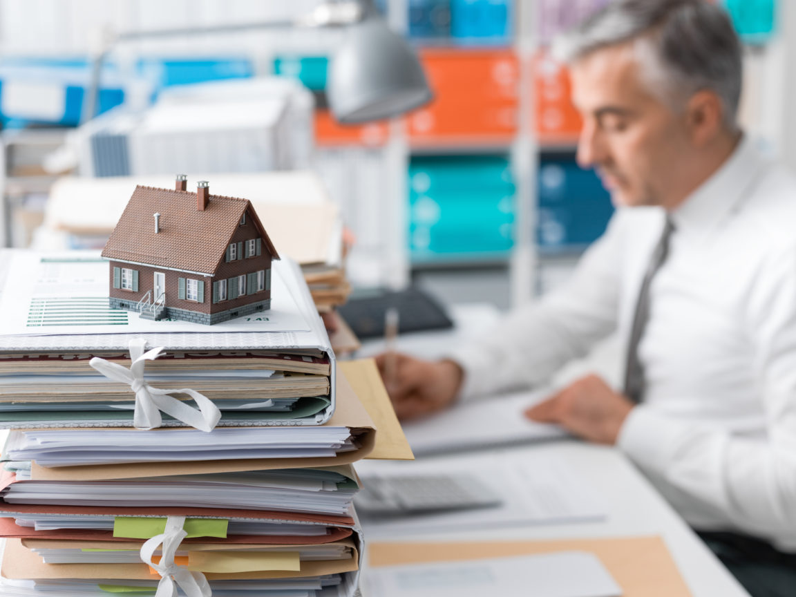 Real estate agent working in the office and piles of paperwork, model house on the foreground and mortgage loan documentation