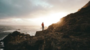 man standing on brown mountain while looking on body of water during daytime