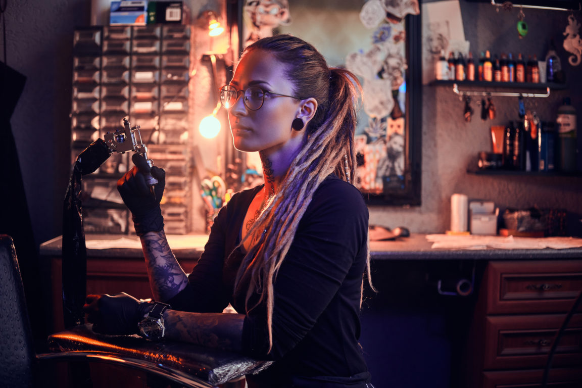 young pretty tattoo master with dreadlocks and glasses is taking short break while holding tattoo machine.