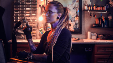 young pretty tattoo master with dreadlocks and glasses is taking short break while holding tattoo machine.