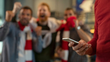 close up of man holding smartphone and betting on the game while supporters celebrating the victory in background. man using mobile in pub during football match. soccer fans cheering for league in pub while man using smart phone to gamble with copy space.