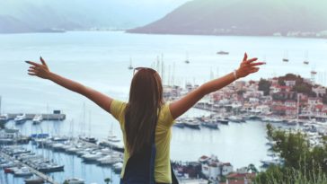 woman raising her hands facing cityscape near body of water