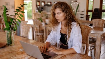 content female customer with long curly hair wearing casual outfit sitting at wooden table with netbook in classic interior restaurant while making online order